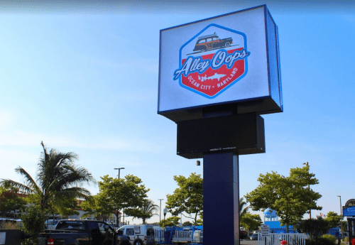 Sign for "Alley Oops" in Ocean City, featuring a retro car design and surrounded by palm trees and blue sky.