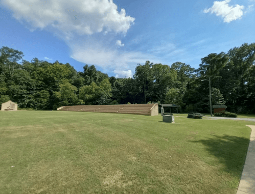 A grassy area with stone seating and trees under a blue sky with clouds.