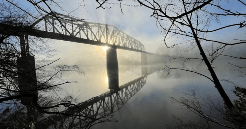 A foggy river scene with a silhouetted bridge reflecting in the water, framed by bare branches.