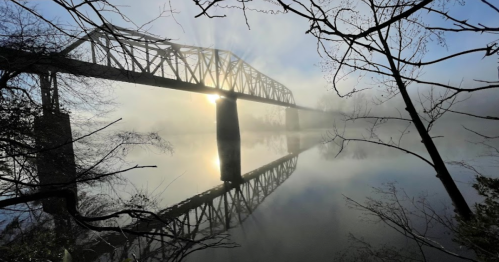 A foggy river scene with a silhouetted bridge reflecting in the water, framed by bare branches.