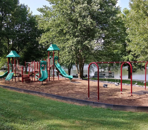 Playground with slides and swings, surrounded by trees and a lake in the background on a sunny day.