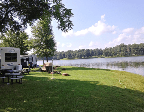 A serene lakeside scene with RVs parked on green grass, surrounded by trees and a calm water view under a blue sky.