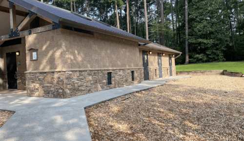 A rustic building with stone accents, surrounded by gravel and trees, set in a natural outdoor environment.