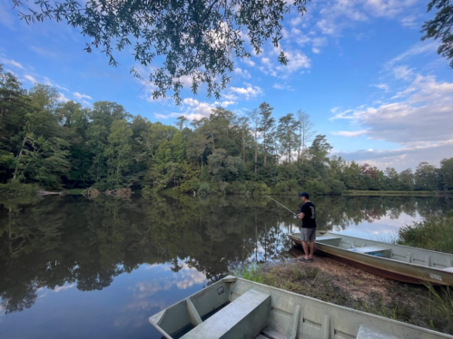 A person fishing by a calm lake surrounded by trees, with two boats in the foreground under a blue sky.
