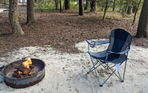 A camping chair beside a fire pit with a small fire, set in a wooded area with sandy ground.