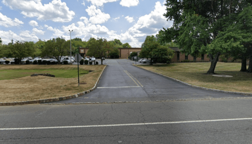 A wide view of a paved road leading to a building, surrounded by trees and grassy areas under a cloudy sky.