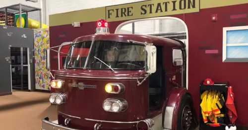 A vintage fire truck parked at a colorful fire station entrance, with firefighter gear hanging nearby.