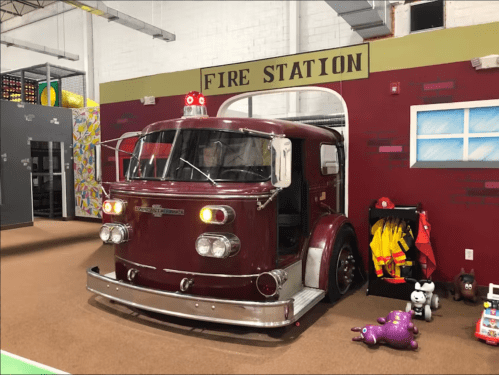 A vintage red fire truck toy inside a play area, with colorful walls and various toys around it.