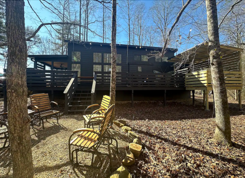 A modern black cabin surrounded by trees, with a deck and seating area on a leaf-covered ground.