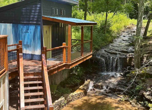 A modern cabin with a blue roof overlooks a small waterfall and stream, surrounded by lush greenery.