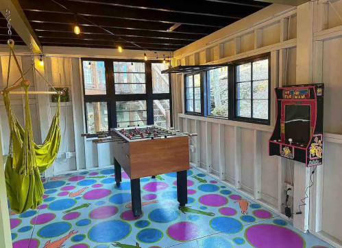 A cozy game room featuring a foosball table, arcade machine, and colorful patterned floor with hanging chairs.