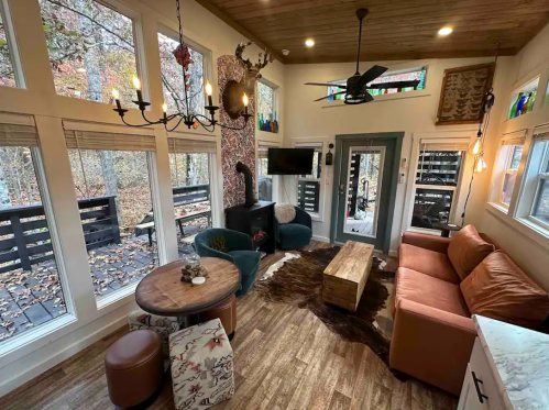 Cozy living room with large windows, a sofa, wooden table, and rustic decor, surrounded by trees outside.