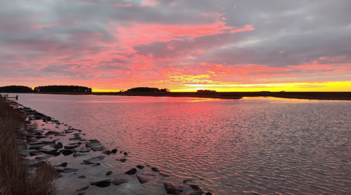 A vibrant sunset over a calm waterway, with colorful clouds reflecting on the surface and rocky shoreline in the foreground.