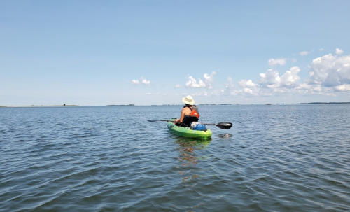 A person in a kayak paddles on a calm water body under a clear blue sky with scattered clouds.