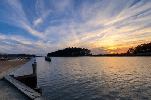 A serene sunset over a calm waterway, with a wooden pier extending into the scene and trees in the distance.