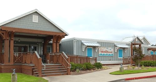 A charming waterfront building with a wooden porch and blue doors, surrounded by greenery and a cloudy sky.