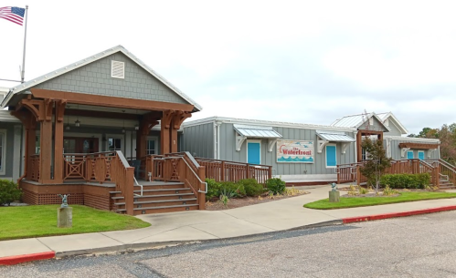 A charming building with a wooden porch, steps, and a banner, surrounded by landscaping and a cloudy sky.