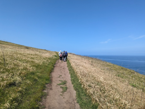 A group of people walking along a dirt path beside a grassy hill, with the ocean visible in the background under a clear blue sky.