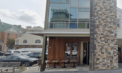 Modern restaurant exterior with large windows, stone accents, and outdoor seating area. Cars parked nearby.