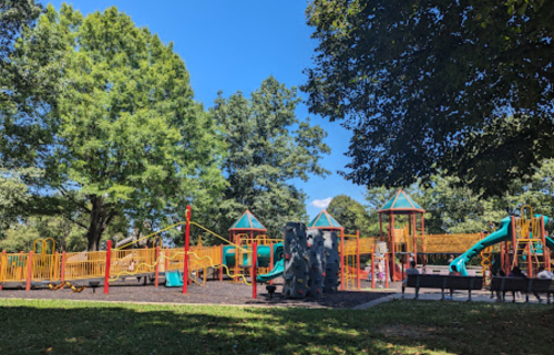 A colorful playground with slides and climbing structures, surrounded by green trees under a clear blue sky.