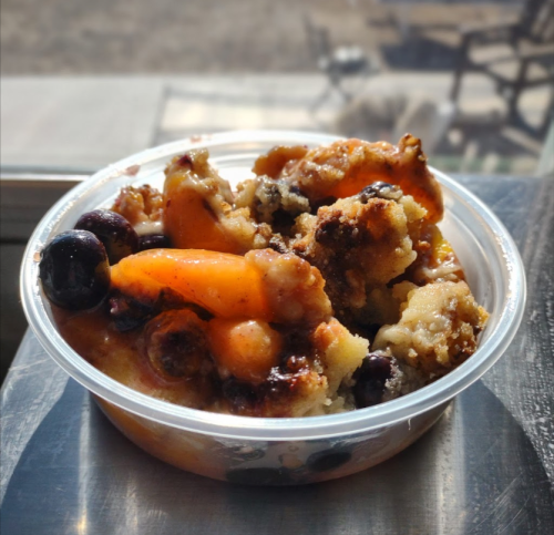 A clear plastic cup filled with a dessert featuring blueberries, orange pieces, and a crumbly topping.
