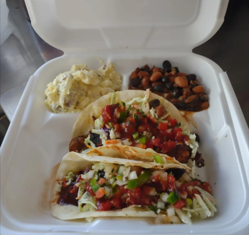 A takeout container with two tacos topped with salsa, alongside sides of potato salad and mixed beans.