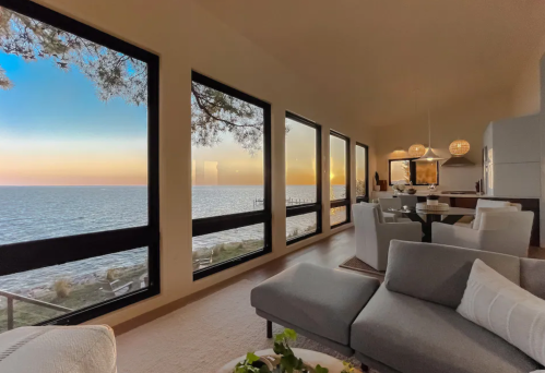 Cozy living space with large windows overlooking a serene sunset by the water. Modern decor and a dining area visible.