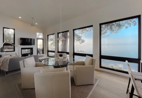 Modern living room with a glass dining table, white chairs, and large windows overlooking a serene lake view.