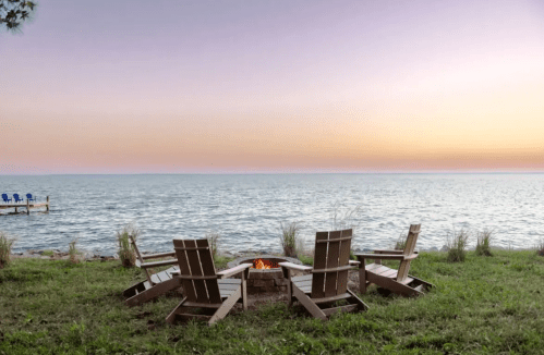 A serene lakeside scene with four wooden chairs around a fire pit, overlooking calm water at sunset.