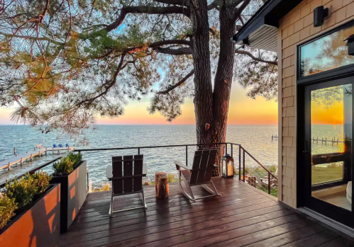 A serene deck with two chairs overlooking a calm lake at sunset, framed by a large tree and colorful sky.