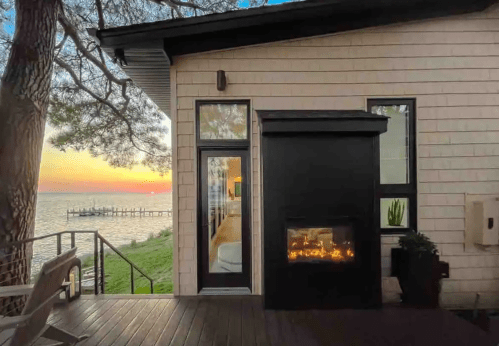 A cozy outdoor space with a fireplace, overlooking a sunset by the water and a pier in the distance.