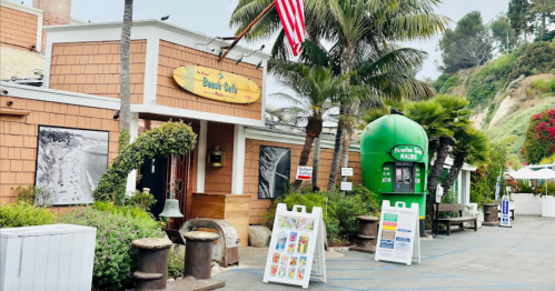 A cozy café with a wooden exterior, palm trees, and an American flag, featuring outdoor seating and colorful signage.