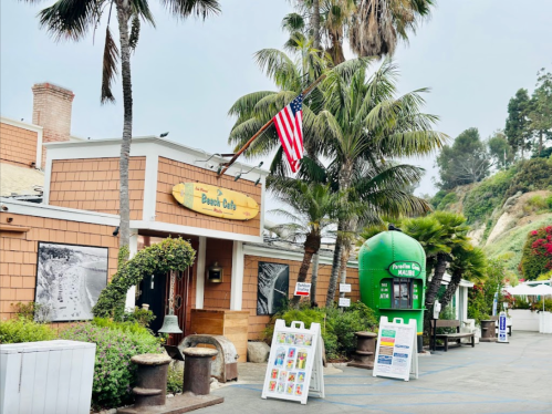 A coastal restaurant with palm trees, an American flag, and colorful signage in a scenic outdoor setting.