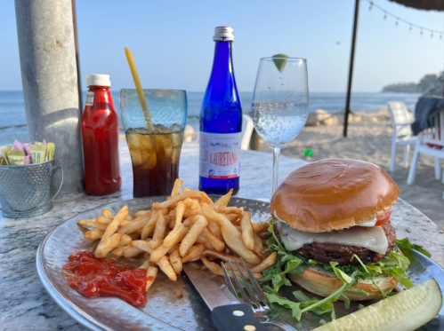 A beachside table with a burger, fries, ketchup, drinks, and a scenic ocean view in the background.