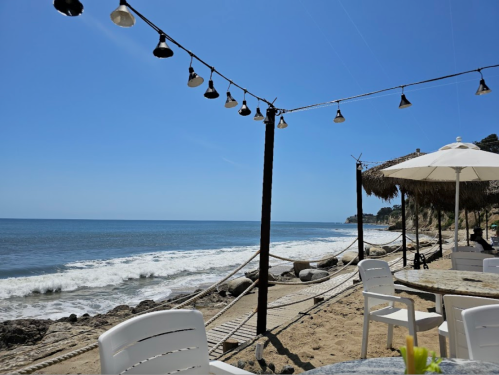 A sunny beach scene with ocean waves, white chairs, and string lights overhead, creating a relaxing atmosphere.