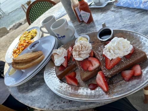 A plate of chocolate waffles topped with strawberries and whipped cream, alongside a side of toast and a colorful breakfast dish.