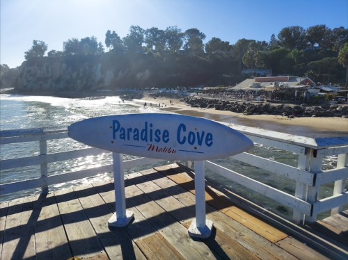 A sign reading "Paradise Cove, Malibu" on a wooden pier overlooking a sandy beach and calm water.