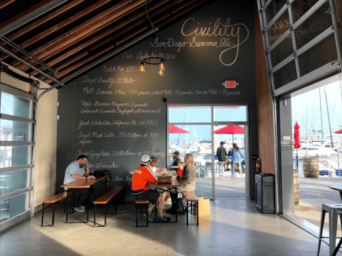 A cozy waterfront brewery with patrons enjoying drinks at a table, large windows, and a chalkboard menu on the wall.