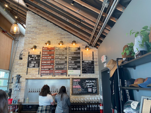 A rustic bar with a brick wall, wooden beams, and a menu board displaying various drinks and snacks. Two people are at the counter.