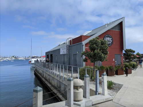 A waterfront brewery building with a red exterior, surrounded by water and boats under a partly cloudy sky.