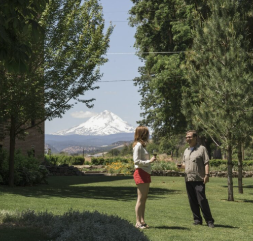 A woman in a white top and red shorts talks to a man in a gray shirt, with a snow-capped mountain in the background.