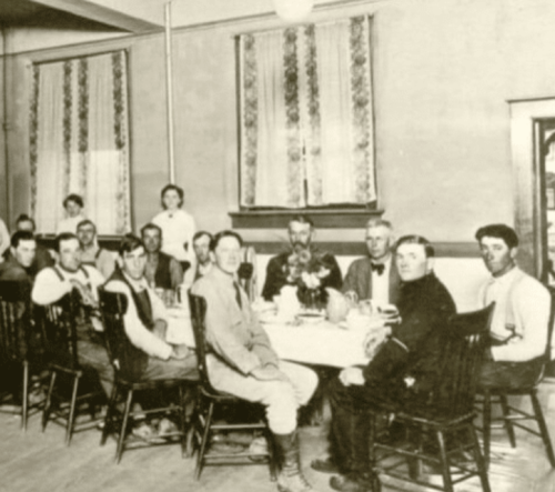 A historical black-and-white photo of a group of men and women seated around a dining table in a rustic setting.