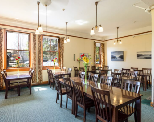 A bright dining room with wooden tables, chairs, and flower centerpieces, featuring large windows and artwork on the walls.