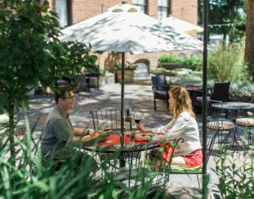 A man and woman sit at a table under an umbrella, enjoying drinks in a sunny outdoor garden setting.