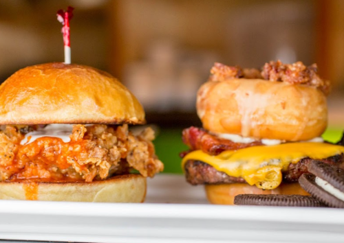 Two gourmet sandwiches on a plate: one with fried chicken and sauce, the other with a burger and donut, topped with cheese.