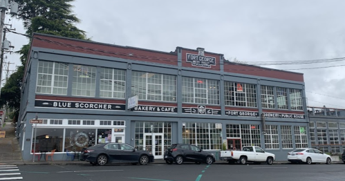 A gray building with large windows, featuring a bakery and café, and a pub, set against a cloudy sky.