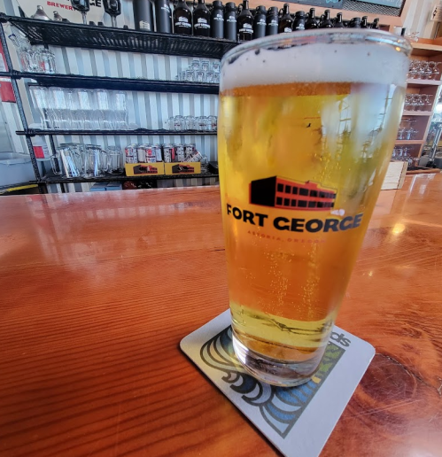 A glass of beer with a Fort George logo sits on a wooden bar, with shelves of glassware in the background.