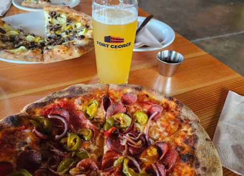 A close-up of a pepperoni and jalapeño pizza with a glass of beer on a wooden table, with another pizza in the background.