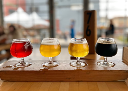 Four small glasses of beer in a wooden flight, featuring red, golden, pale, and dark brews, set in a bright brewery.