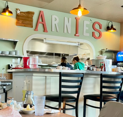 A cozy diner interior with a counter labeled "ARNIE'S," featuring staff and various condiments on the tables.